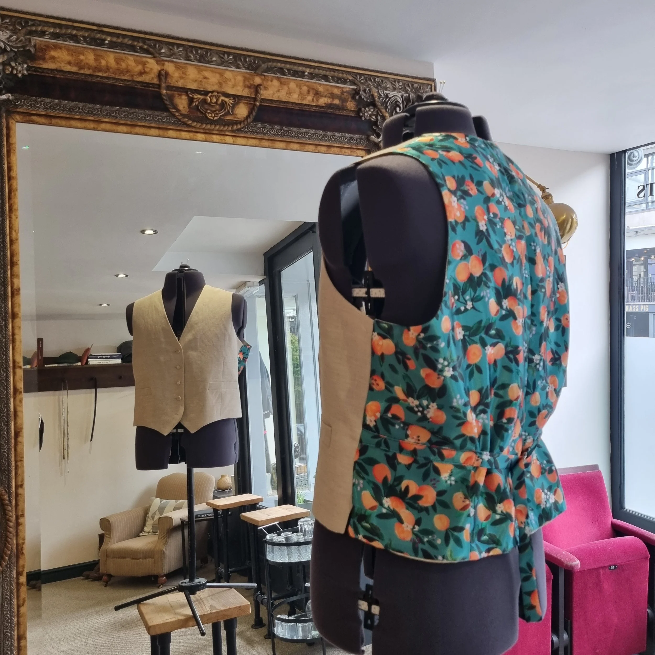 Two mannequins dressed in vintage-style vests and floral tops, positioned in front of a large ornate mirror inside a clothing store.