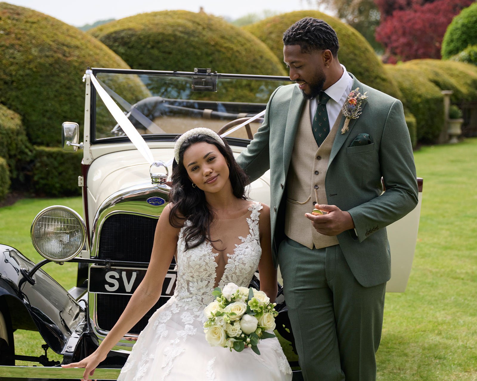 A bride in a lace wedding gown with dark hair holding a bouquet of white roses, and a groom in a green suit with a boutonniere, standing in front of a vintage Ford car decorated with white ribbons in a garden with lush green bushes.