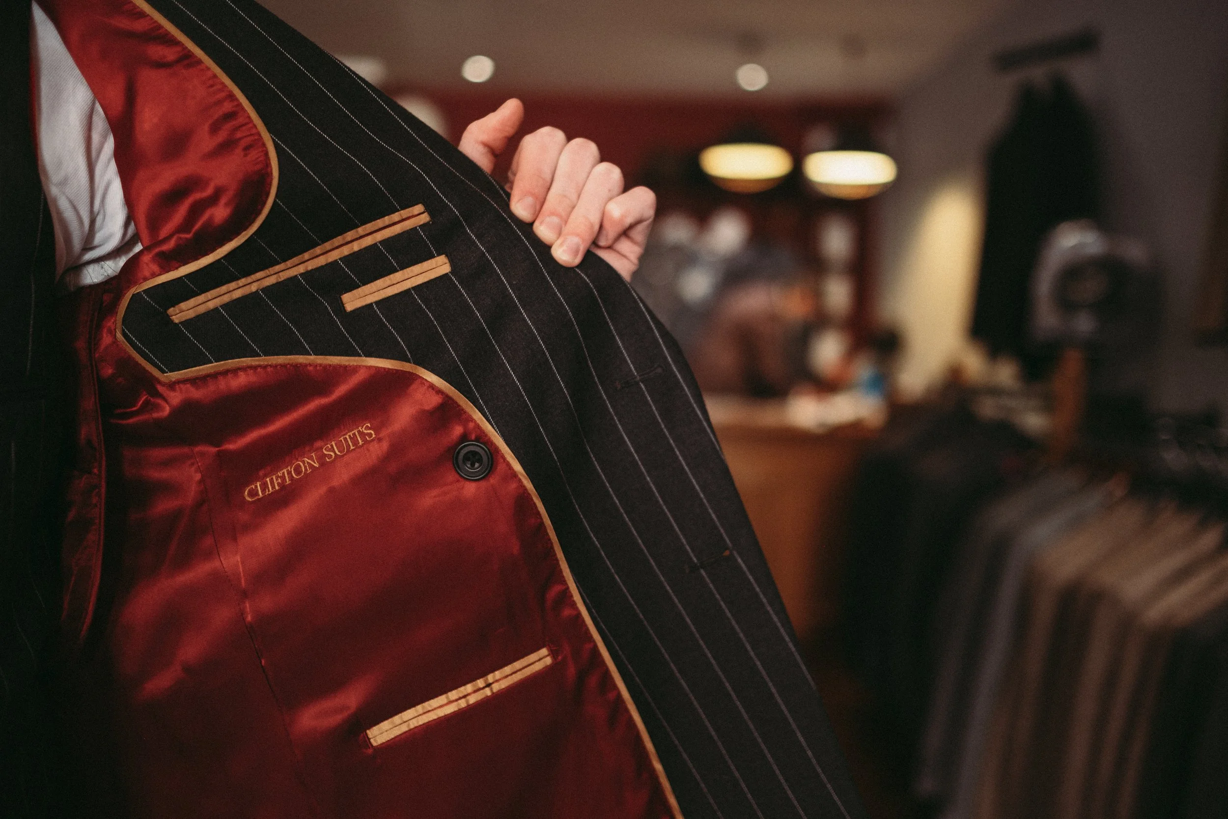 Close-up of a person holding open the inside of a dark pinstripe suit jacket with red lining and the words 'Clifton Suits' embroidered inside.