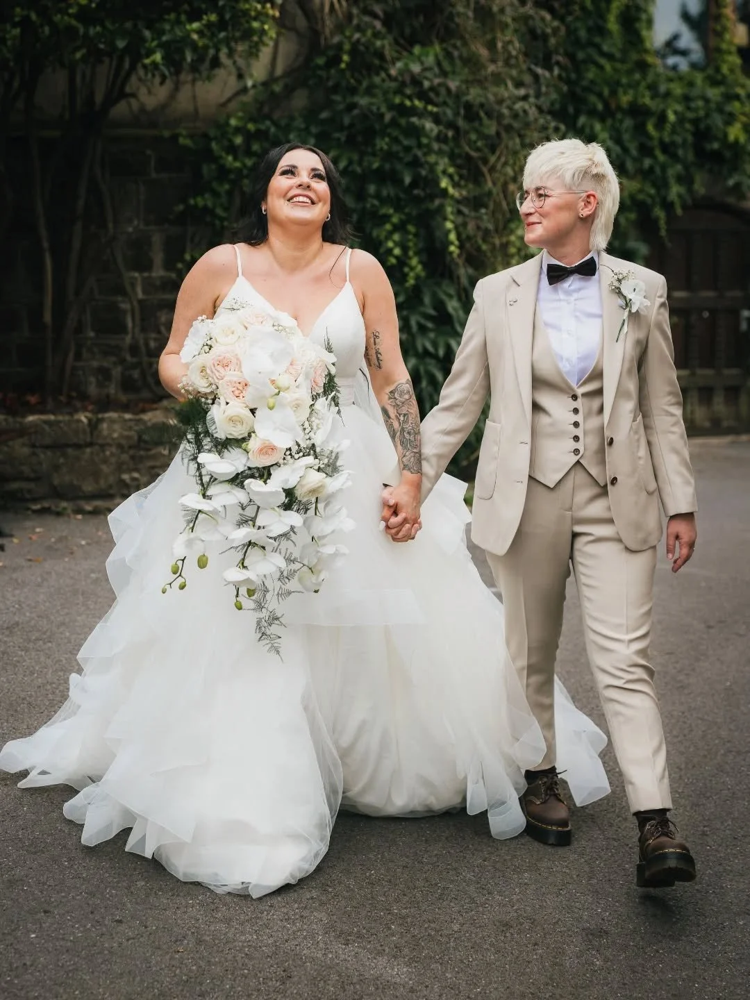 A same-sex couple holding hands and walking outdoors on a wedding day. One person is wearing a white dress and holding a large floral bouquet, the other is wearing a beige suit with a bow tie, both are smiling and looking at each other.