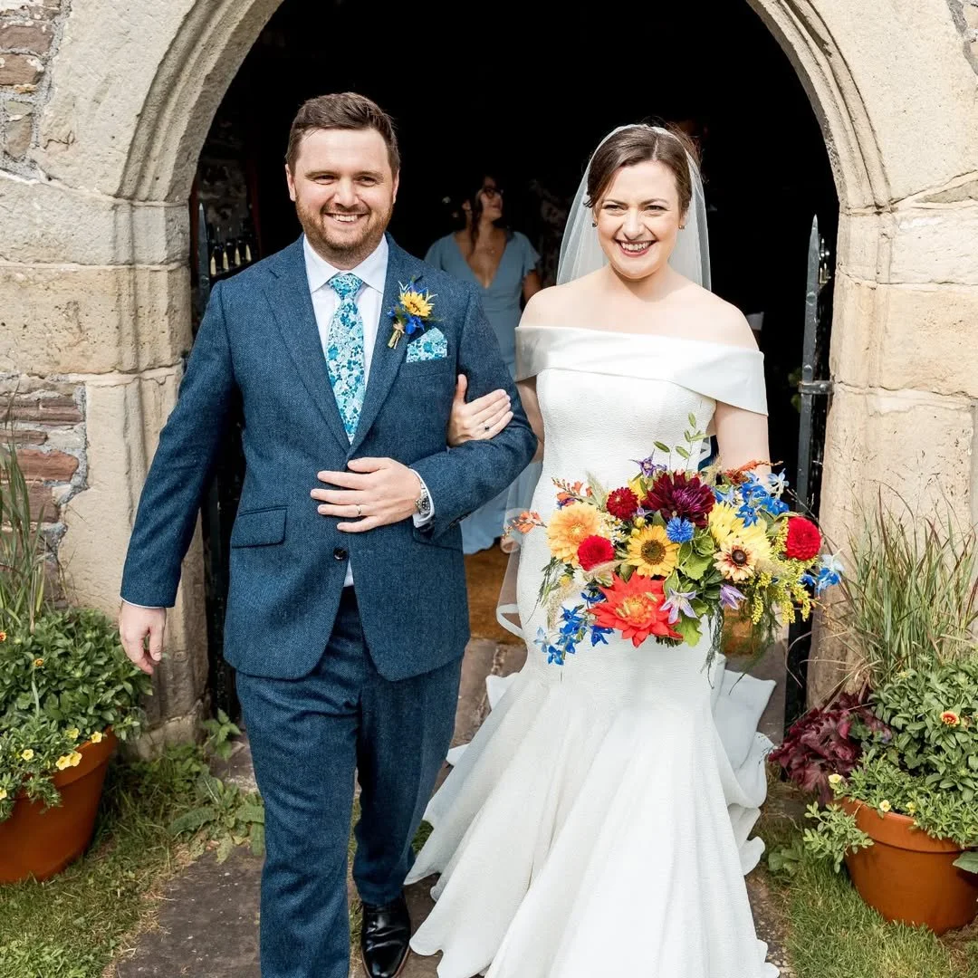 A bride and groom smiling as they walk out of a stone archway, holding hands, with the bride carrying a large colorful bouquet of flowers.