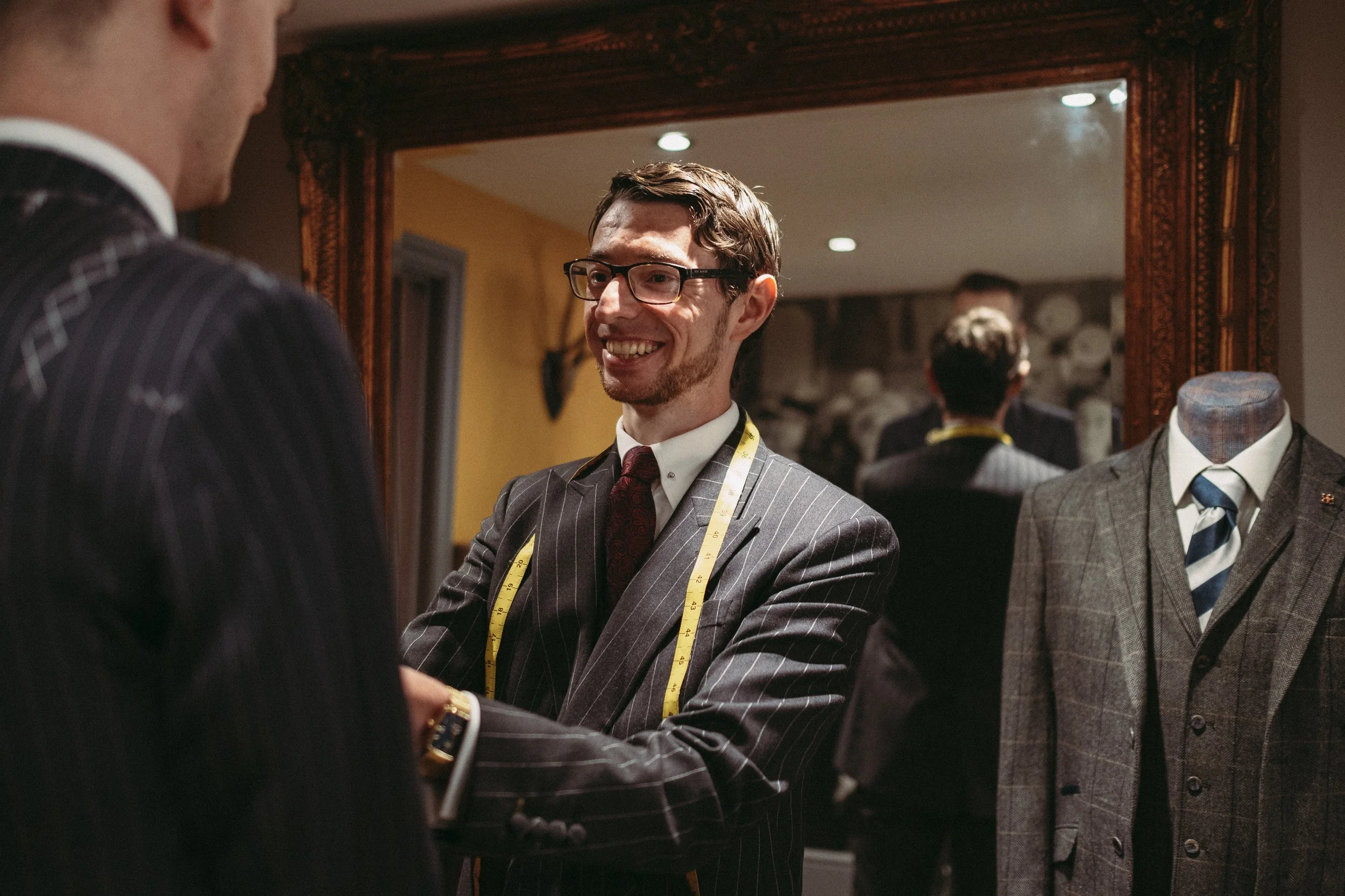 A tailor or clothing designer in a pinstripe suit smiling and shaking hands with a customer in a suit, measuring tape around his neck, in a tailor shop with a mirror and a mannequin dressed in a plaid suit and tie.