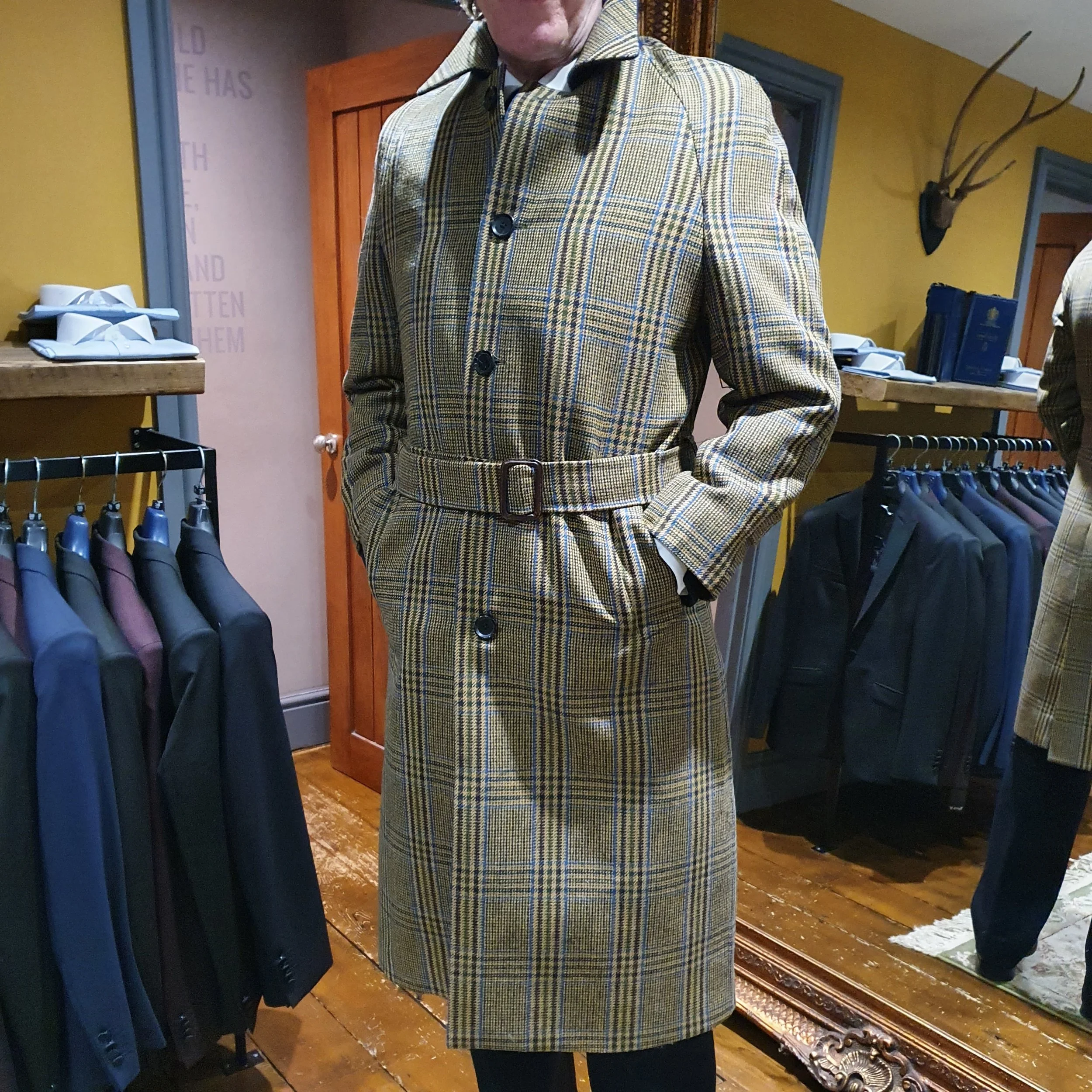 A person wearing a yellow plaid trench coat with a belt, standing inside a clothing store with suits hanging on racks.