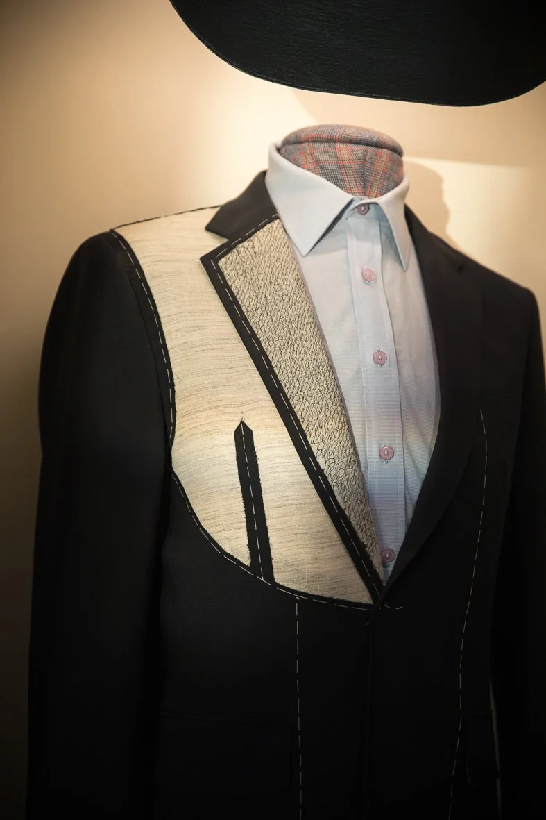 A mannequin dressed in a black blazer with white stitched lines and a white button-up shirt, featuring artistic stitching designs on the chest area, with a plaid inner collar and a black hat.