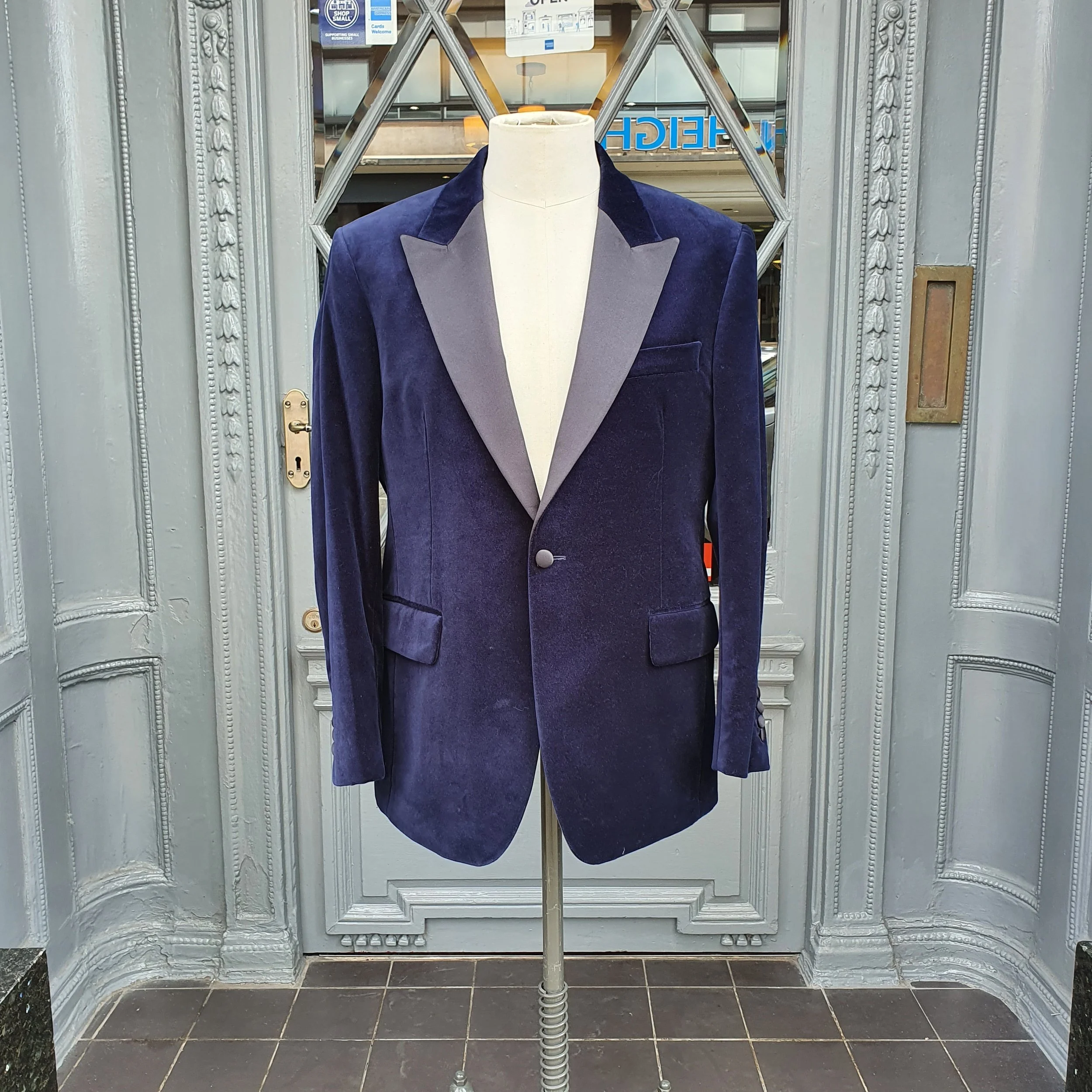 A vintage blue velvet tuxedo jacket with a contrasting dark gray lapel, displayed on a mannequin outside a shop.