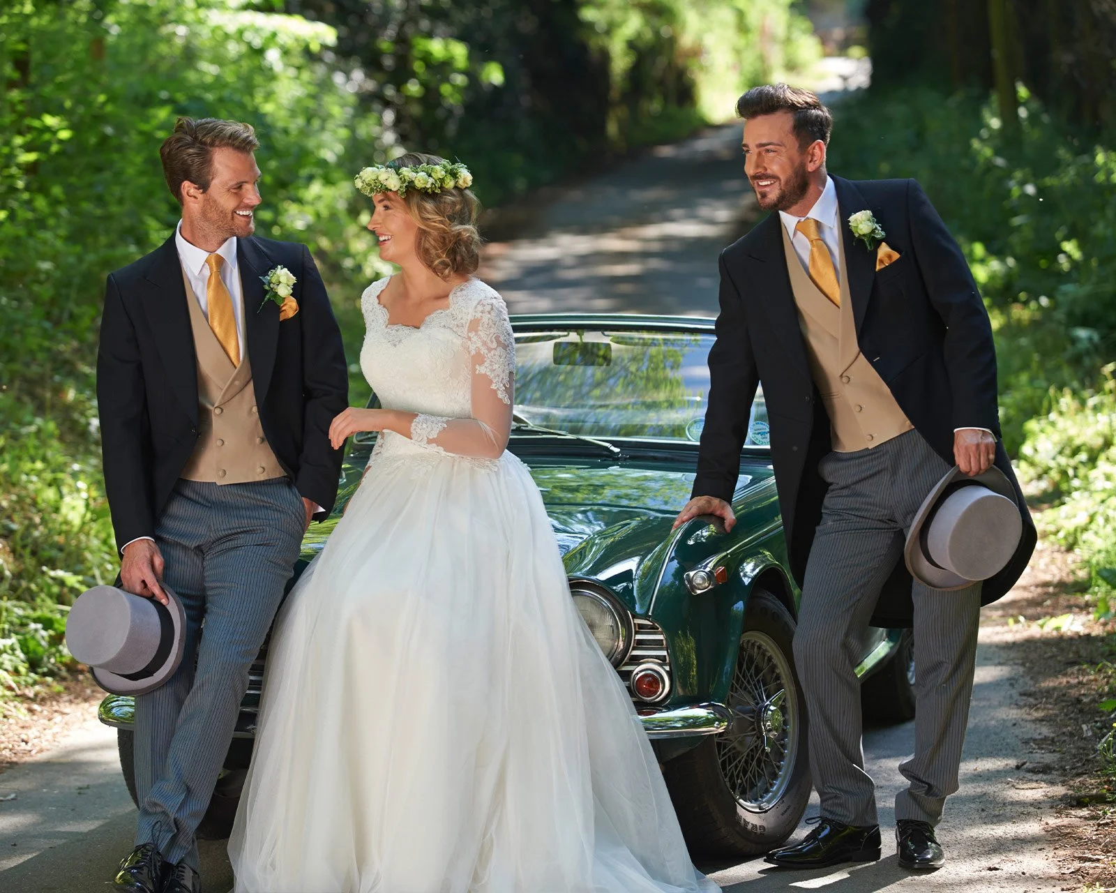 A bride in a white wedding dress and two groomsmen in tuxedos with striped pants and vests, standing by a classic black car on a forested dirt road, all smiling and holding hats, during a wedding photoshoot.