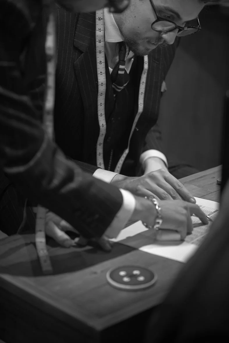 Close-up of a man in a suit with a measuring tape around his neck, working on paperwork at a desk. Another person is assisting, pointing at the paper, in a professional setting.