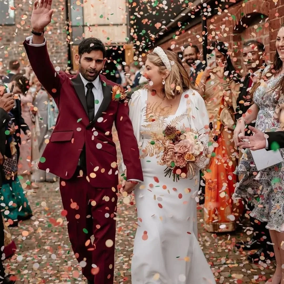 Happy wedding celebration with a bride and groom walking through colorful confetti, surrounded by guests outdoors.