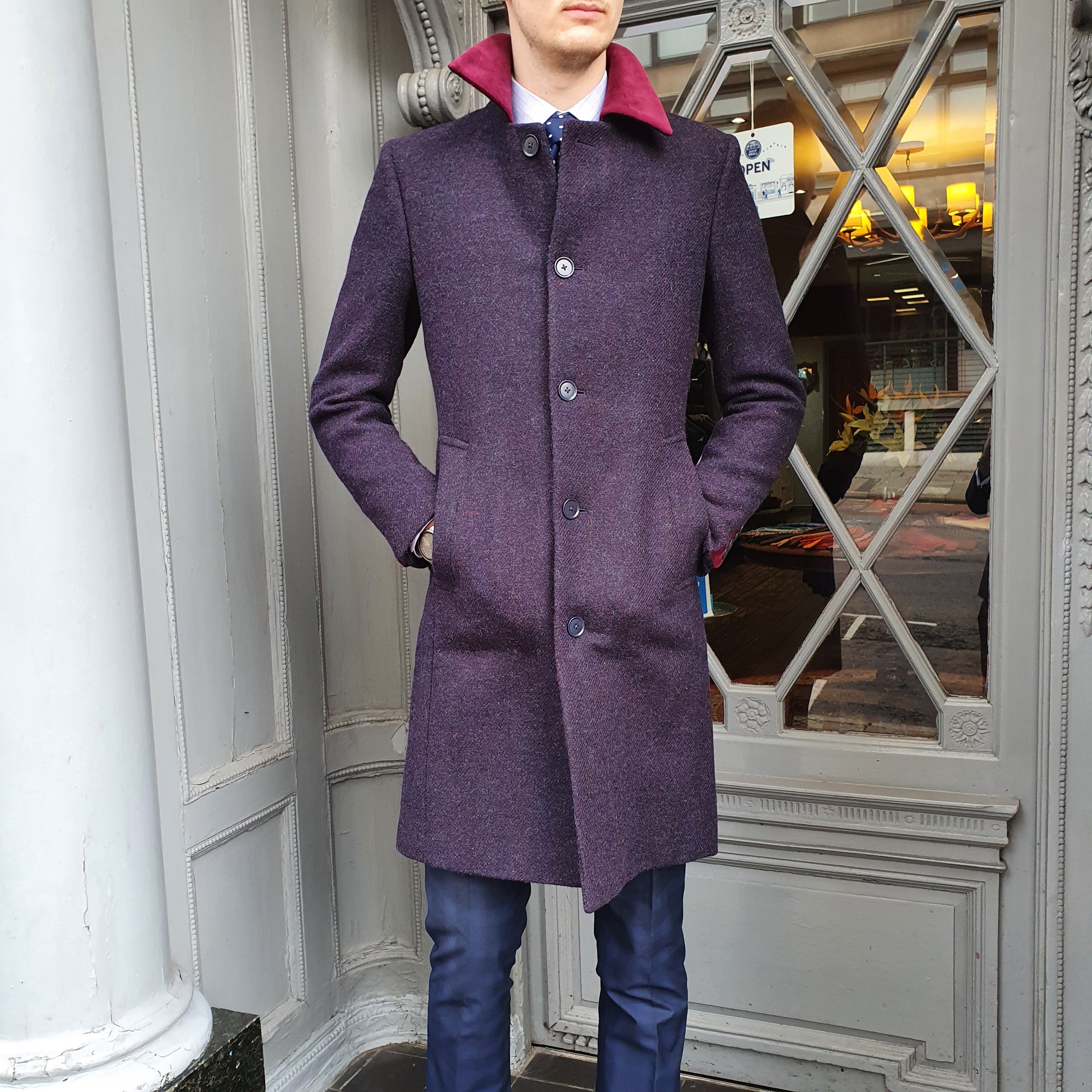 A man wearing a long purple coat with a red collar, white shirt, blue tie, and dark pants stands with his hands in his coat pockets in front of a decorative door and window.