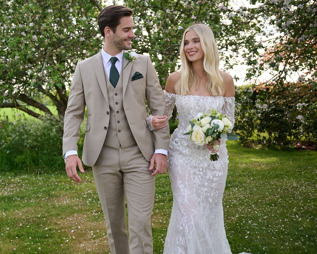 A couple dressed in wedding attire walking outdoors on a sunny day with trees and grass in the background.