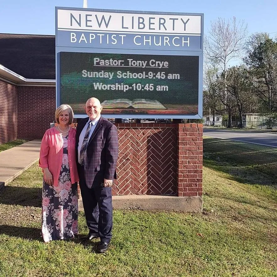 A man and woman standing outside of New Liberty Baptist Church in front of a large sign displaying church information and service times.