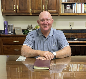 Smiling man sitting at a table in a kitchen, with a book and papers in front of him.