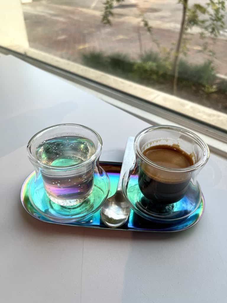  Duas xícaras de vidro, tamanho pequeno, apoiadas em pires furta-cor e de forma alongada. Na xícara da esquerda, há água com gás. Na xícara da direita, uma dose de café espresso. Há uma colher pequena no meio das xícaras. 