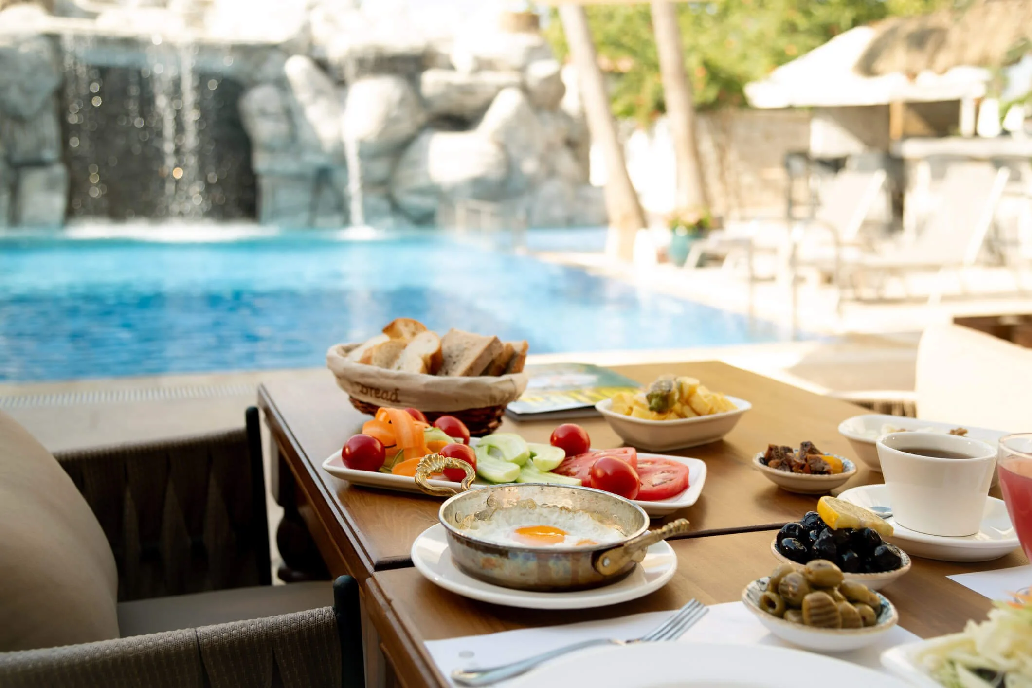 Poolside hotel breakfast featuring fresh fruit, eggs, bread and coffee overlooking an outdoor swimming pool.