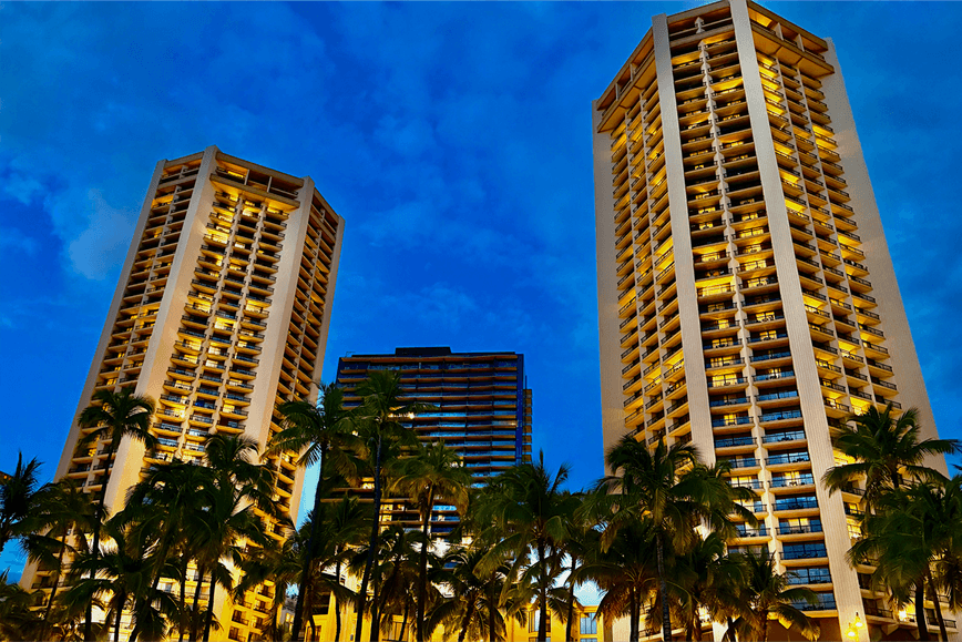 Illuminated luxury hotel towers framed by tropical palm trees against a deep blue evening sky.