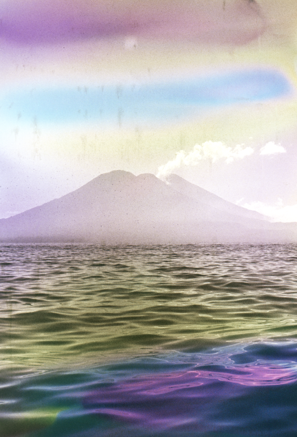 A mountain in the distance across a body of water, with clouds near its peak and rainbow-colored streaks in the sky.