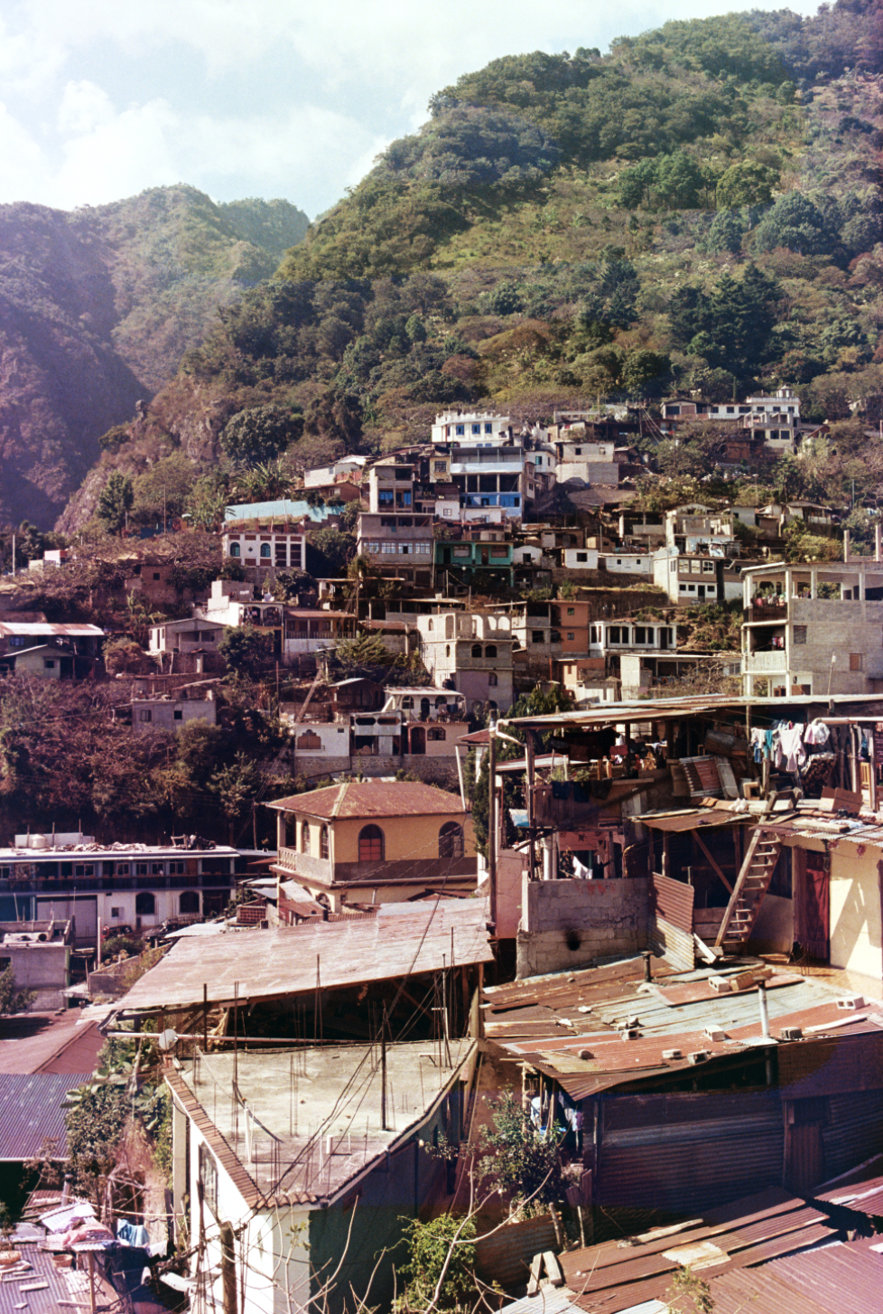 Hilly hillside neighborhood with a mix of colorful houses and apartments stacked on slopes, surrounded by green trees and mountains in the background.