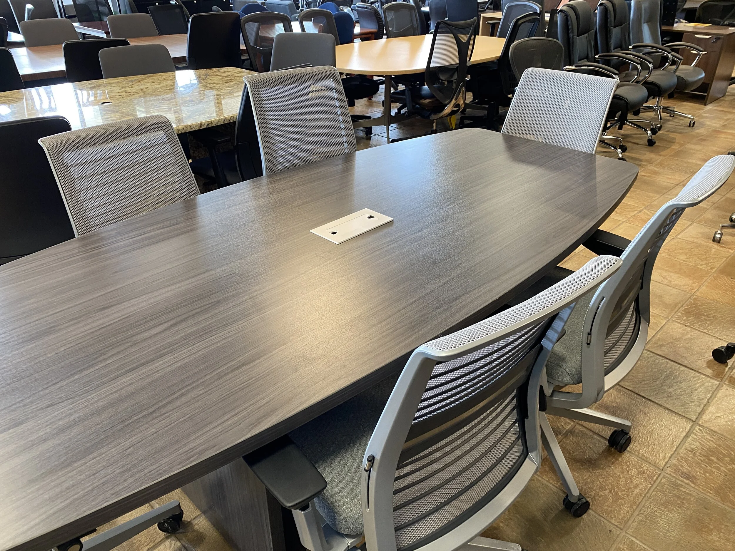 A conference room with a long wooden table and multiple office chairs around it, featuring a power outlet on the table, with additional chairs and tables visible in the background.