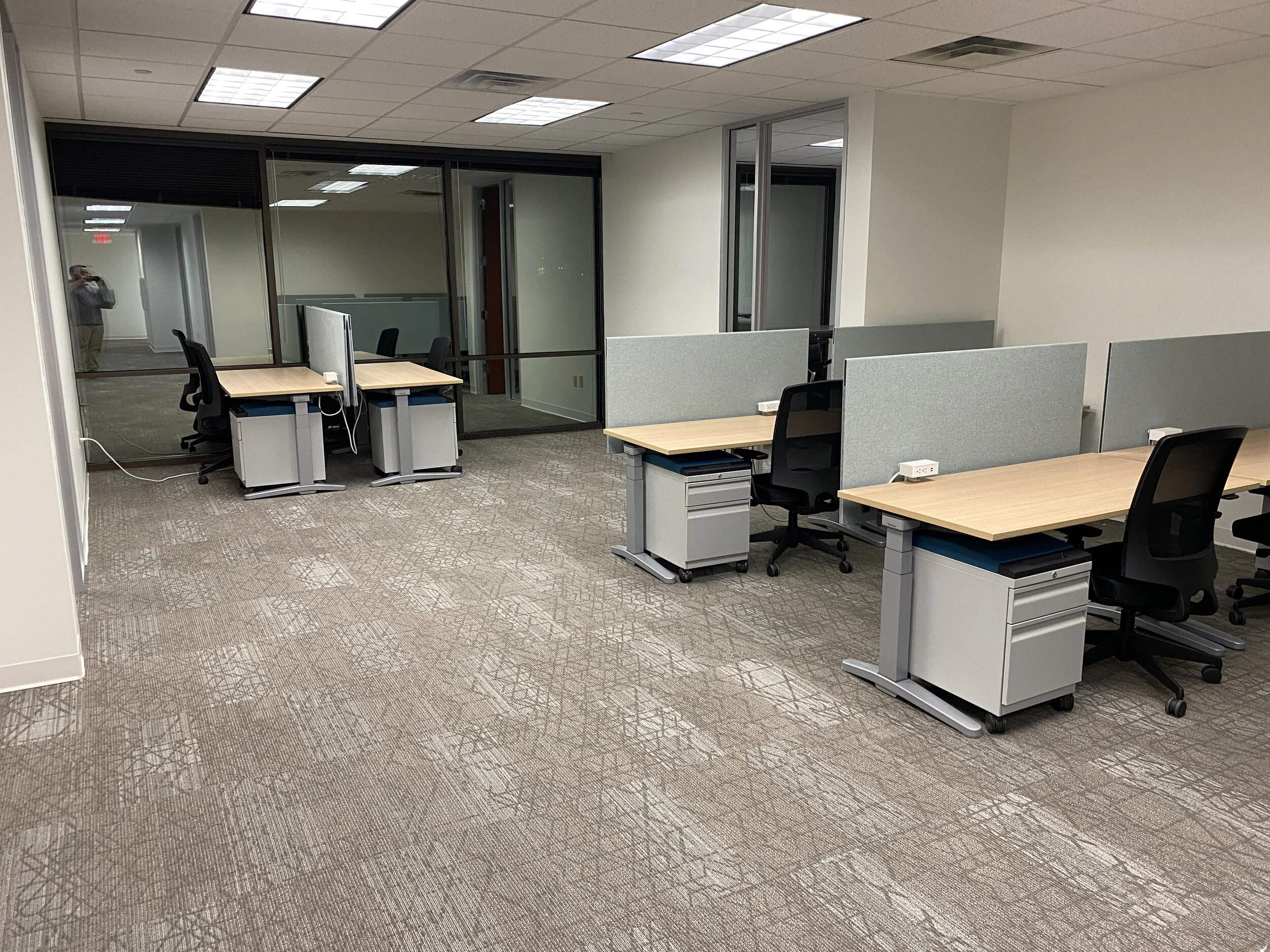 Empty office space with four desks, black chairs, and grey partition panels, with glass-walled conference room in the background. Overhead fluorescent lighting and beige carpet flooring.