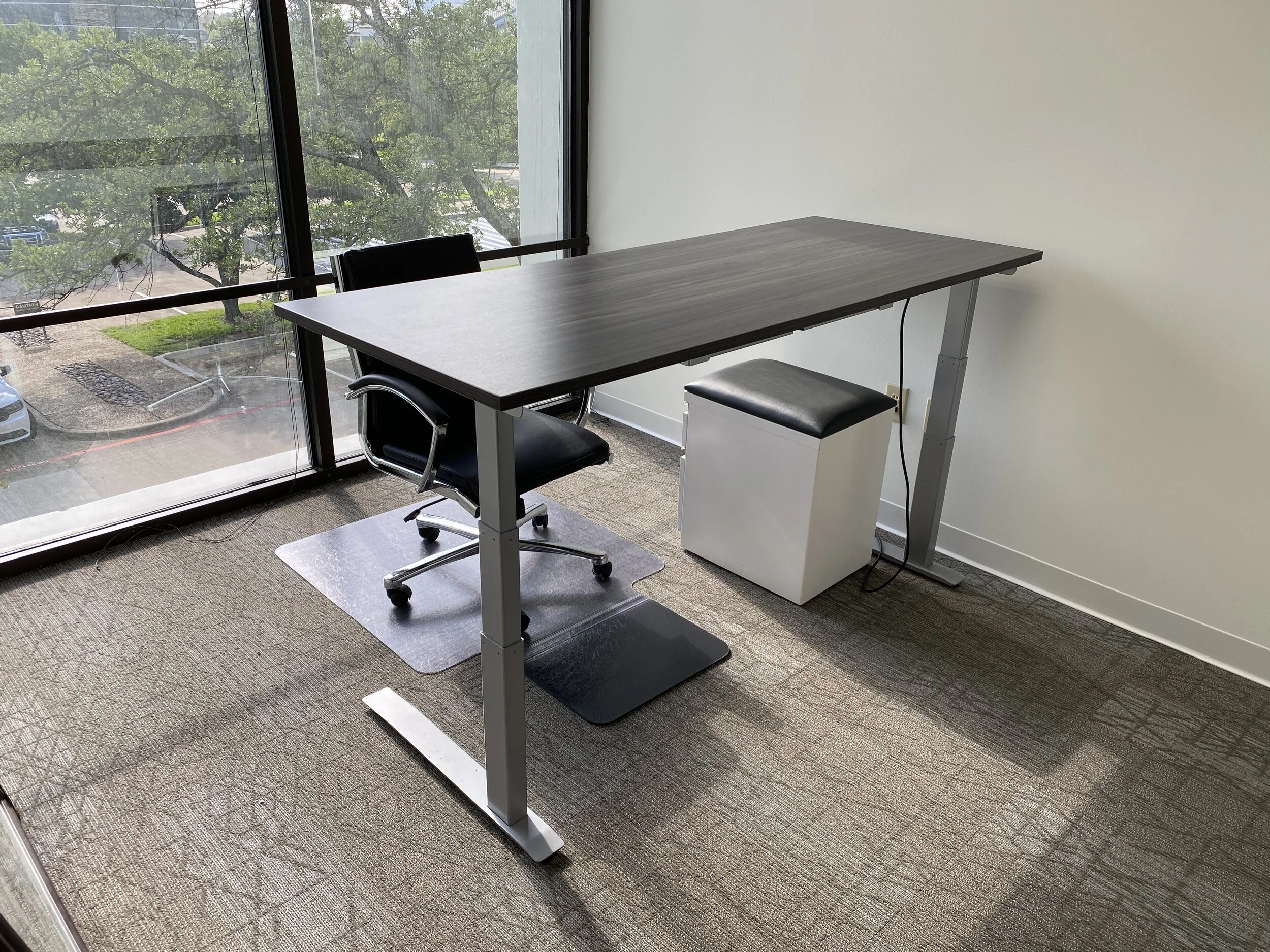 Office workspace with a dark wood desk, a black rolling chair, a black cushioned stool, a white trash bin, and a large window with a view of parked cars and trees.