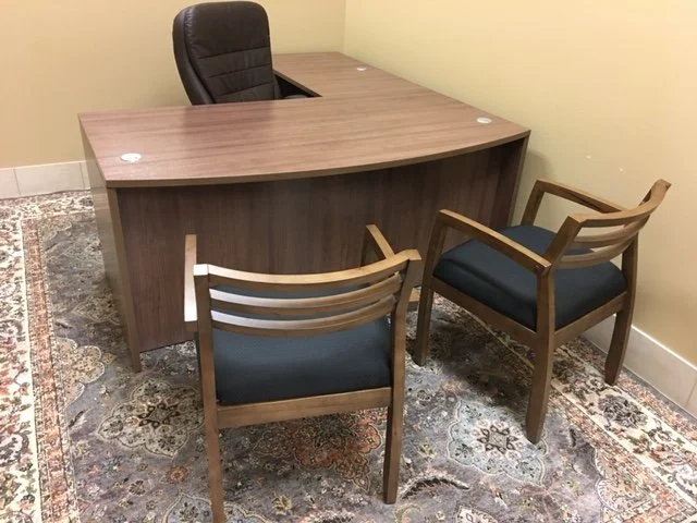An office with a wooden desk and two wooden chairs with dark cushions on an ornate area rug.