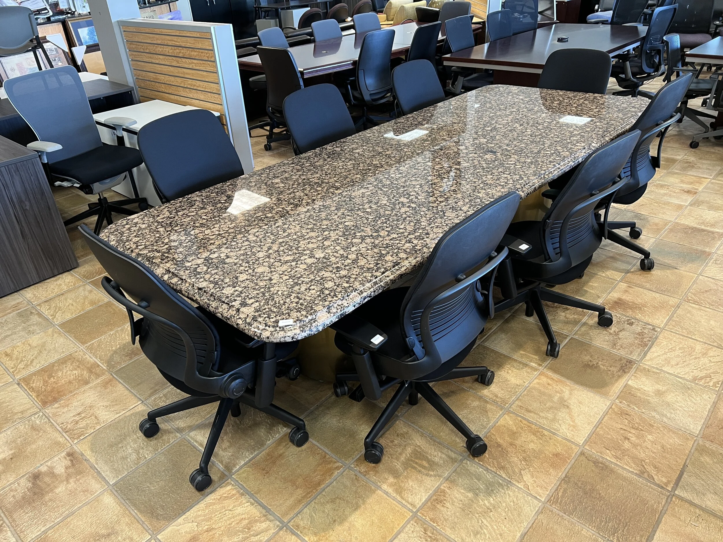 A large rectangular conference table with a granite top surrounded by black office chairs on a tiled floor, with additional office furniture and chairs in the background.