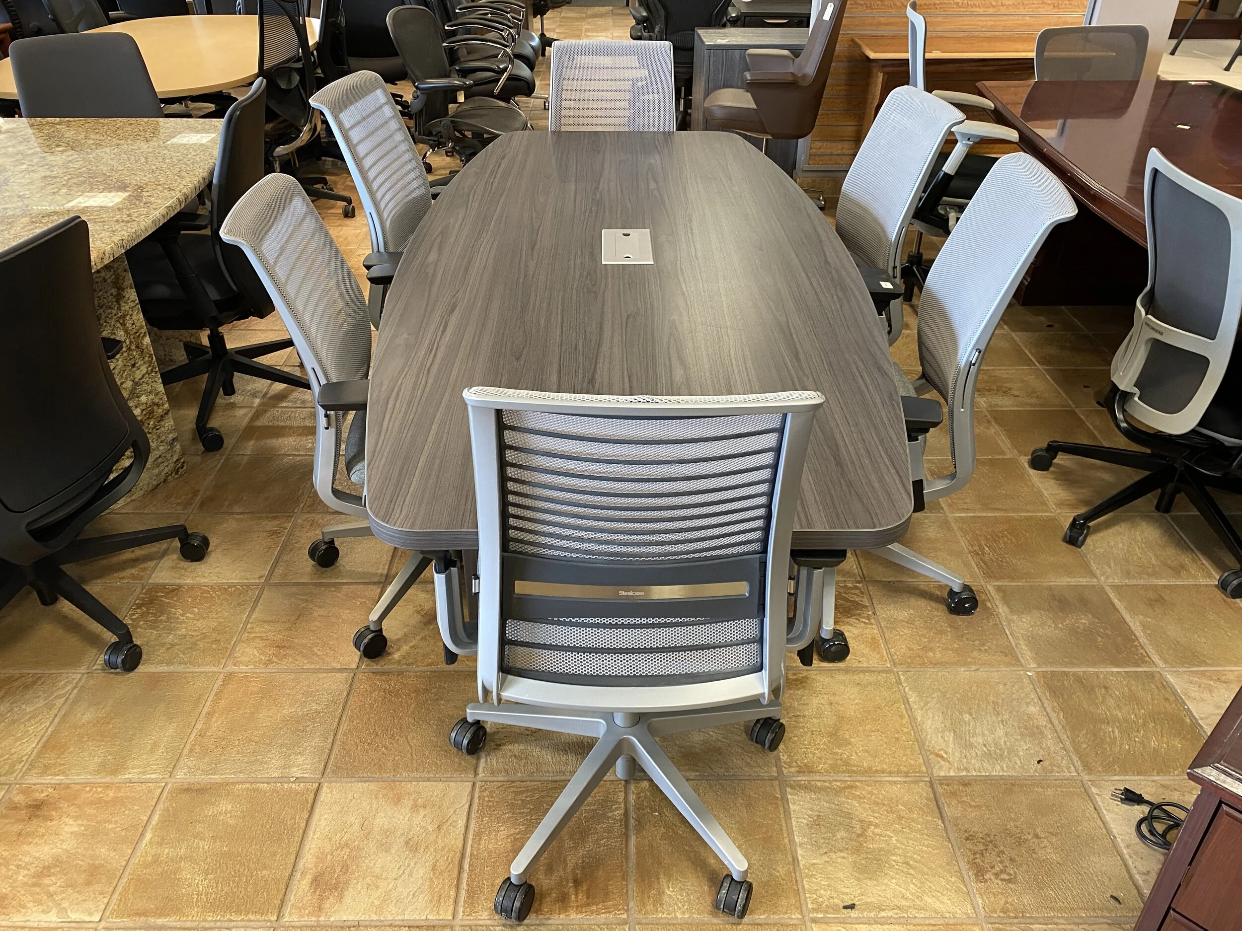 An empty conference room with a large, oval, gray woodgrain table surrounded by nine mesh office chairs with black and gray colors, on a brown tiled floor.