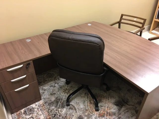Office desk with a black swivel chair, a wooden filing cabinet, and a wooden chair in the background.