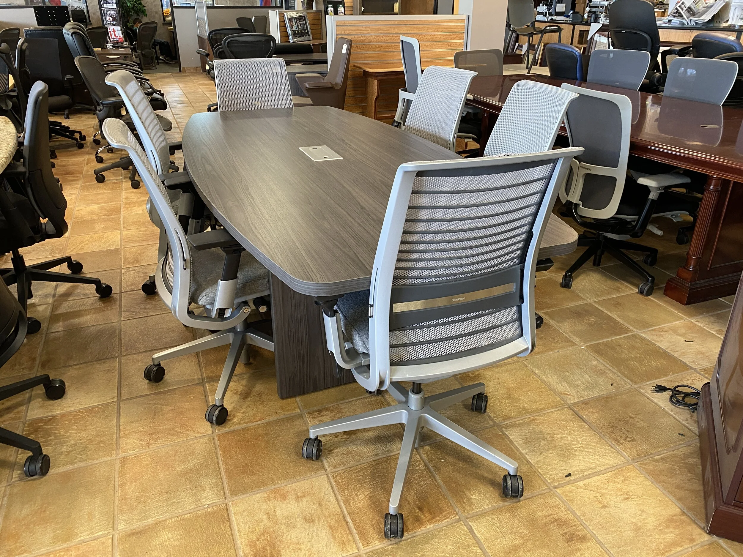 A conference room with a large oval table surrounded by multiple office chairs, some with mesh backs and others with fabric, on a tiled floor.