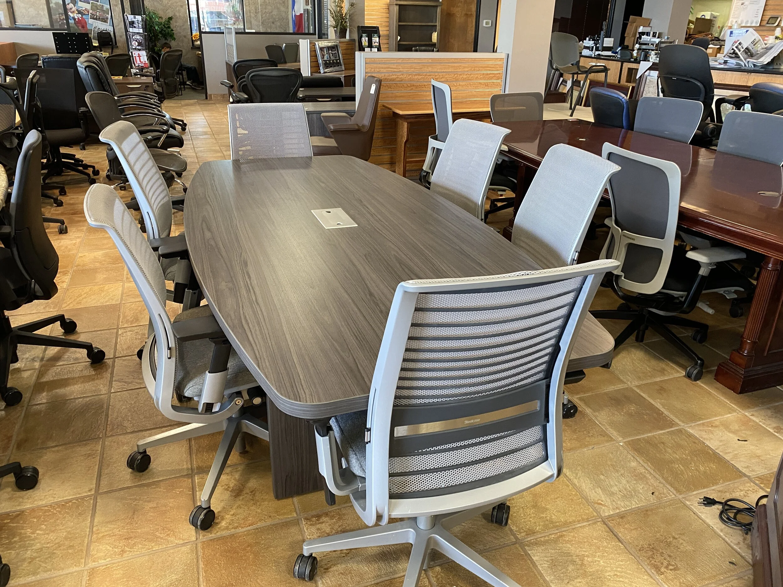A conference room with a large oval wooden table surrounded by multiple office chairs, with additional desks and chairs in the background.