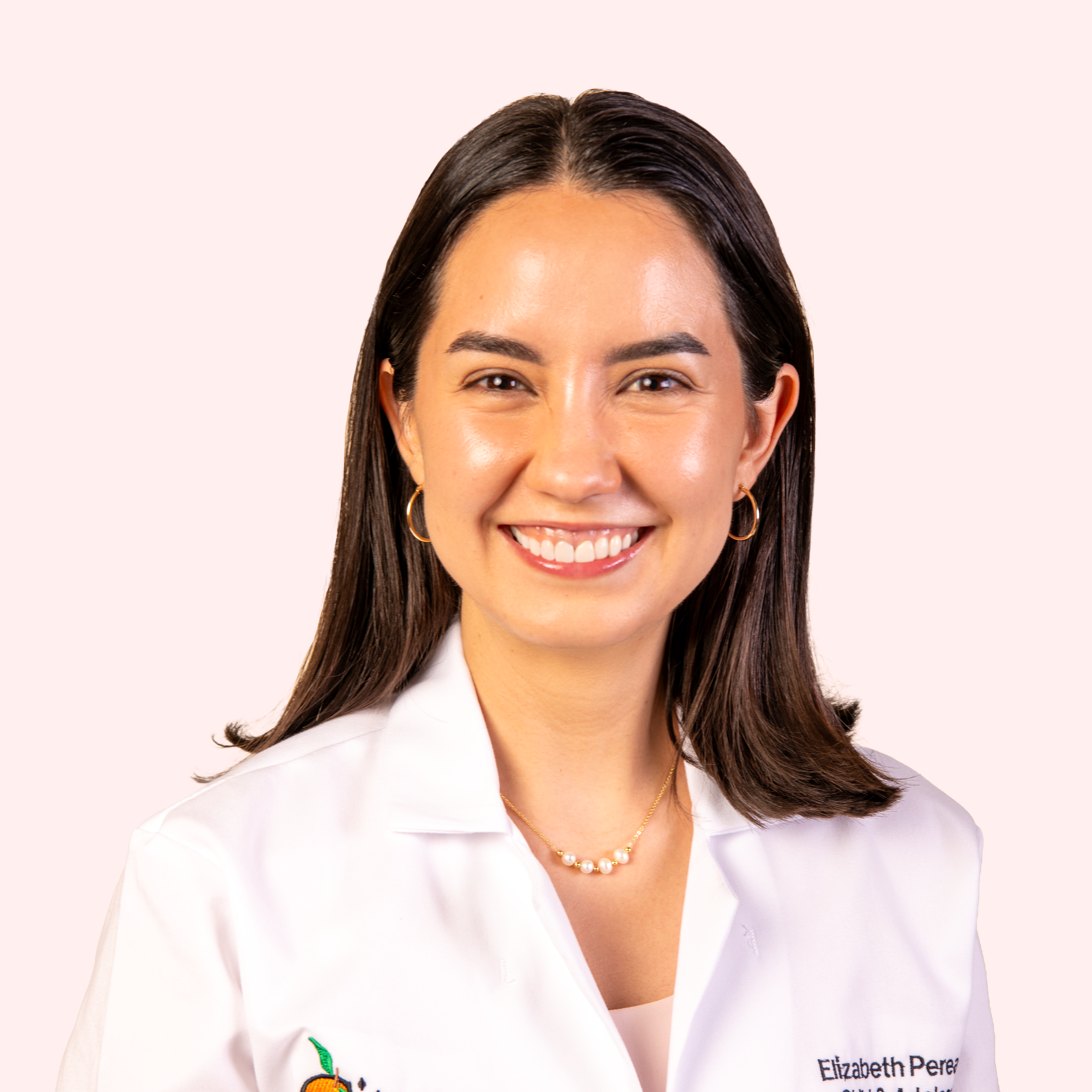 A woman with long, dark hair and a bright smile wearing a white medical coat with text that says 'Beth Perea Psychiatry' on it, standing against a plain light background.