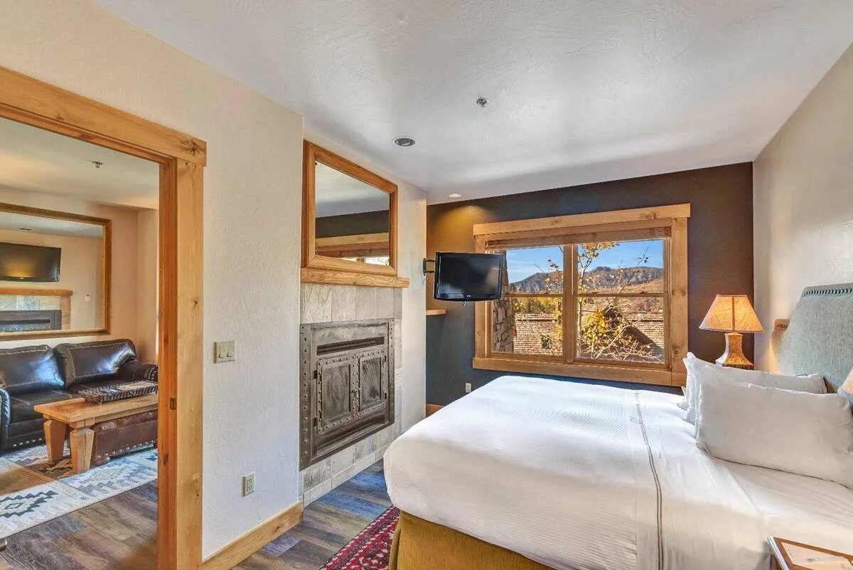 Hotel room with a bed, window view of trees and mountains, wall-mounted TV, bedside lamp, fireplace, and a door leading to a living area with a leather sofa.