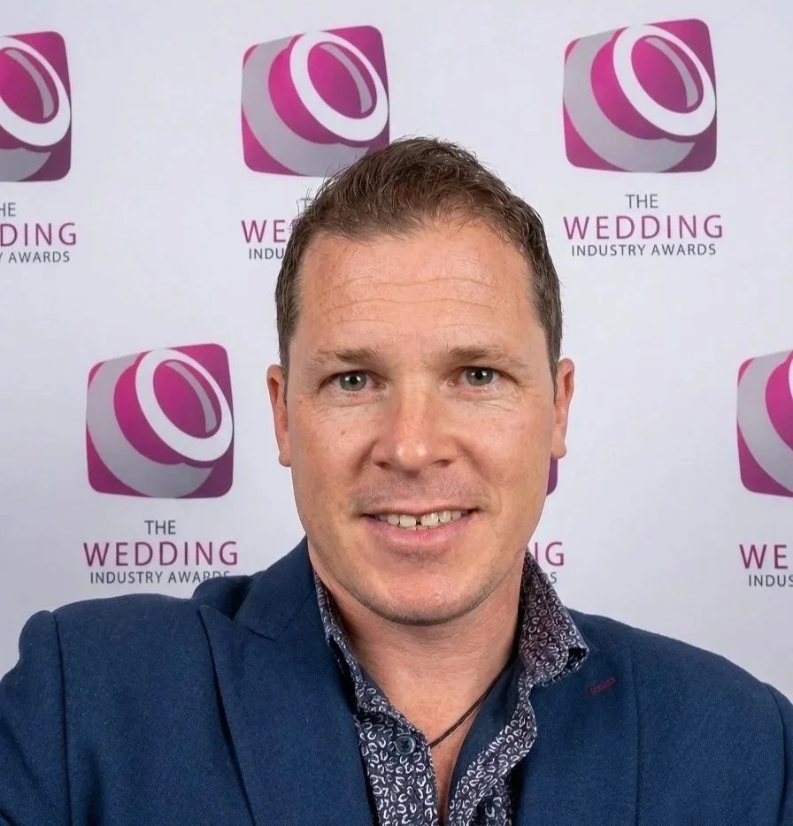 Chris Morris, award winning wedding pianist, smiling at a photo event with a backdrop that says 'The Wedding Industry Awards' and features a pink and purple logo.