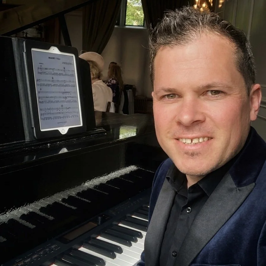 A man is smiling and sitting next to a black piano with an electronic tablet on a music stand displaying sheet music. Two women are visible in the background, sitting at a table in a room with windows, curtains, and some greenery outside.