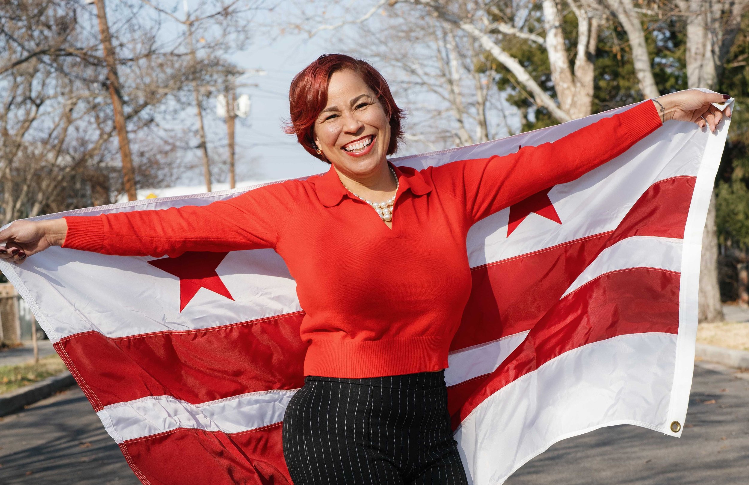 A woman with short red hair and a bright smile holding out a large American flag outdoors during daytime. She's wearing a red top with pinstripe black pants and a pearl necklace. The background shows leafless trees and a clear sky.