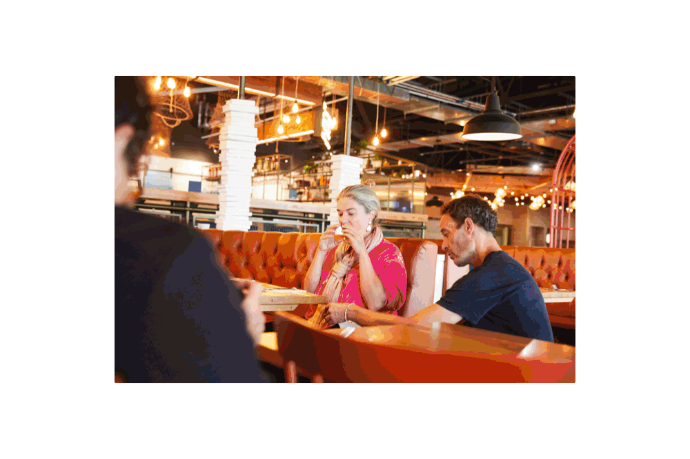A woman and a man sitting at a restaurant table, with the woman about to eat and the man looking down, in a warmly lit restaurant with orange upholstered seating and decorative lighting.