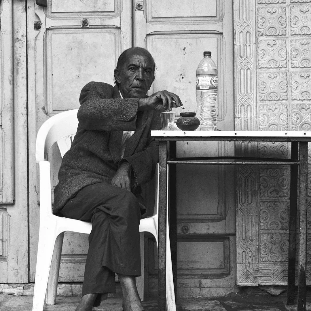 An elderly man sitting on a white plastic chair at a table outside, with a large bottled water, a glass, and a small pot on the table. The background features a detailed carved wall and wooden door.