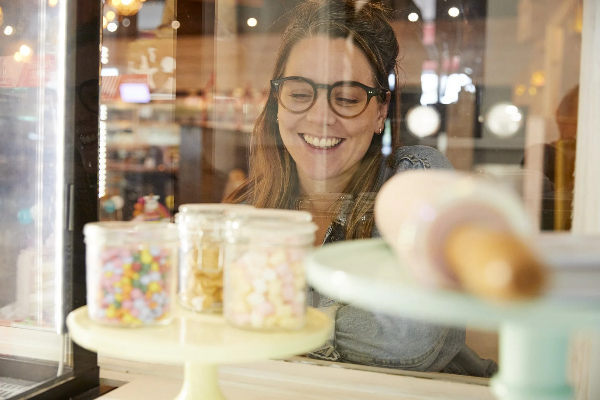 A woman with glasses smiling as she looks inside a bakery window featuring jars filled with colorful candies and desserts.