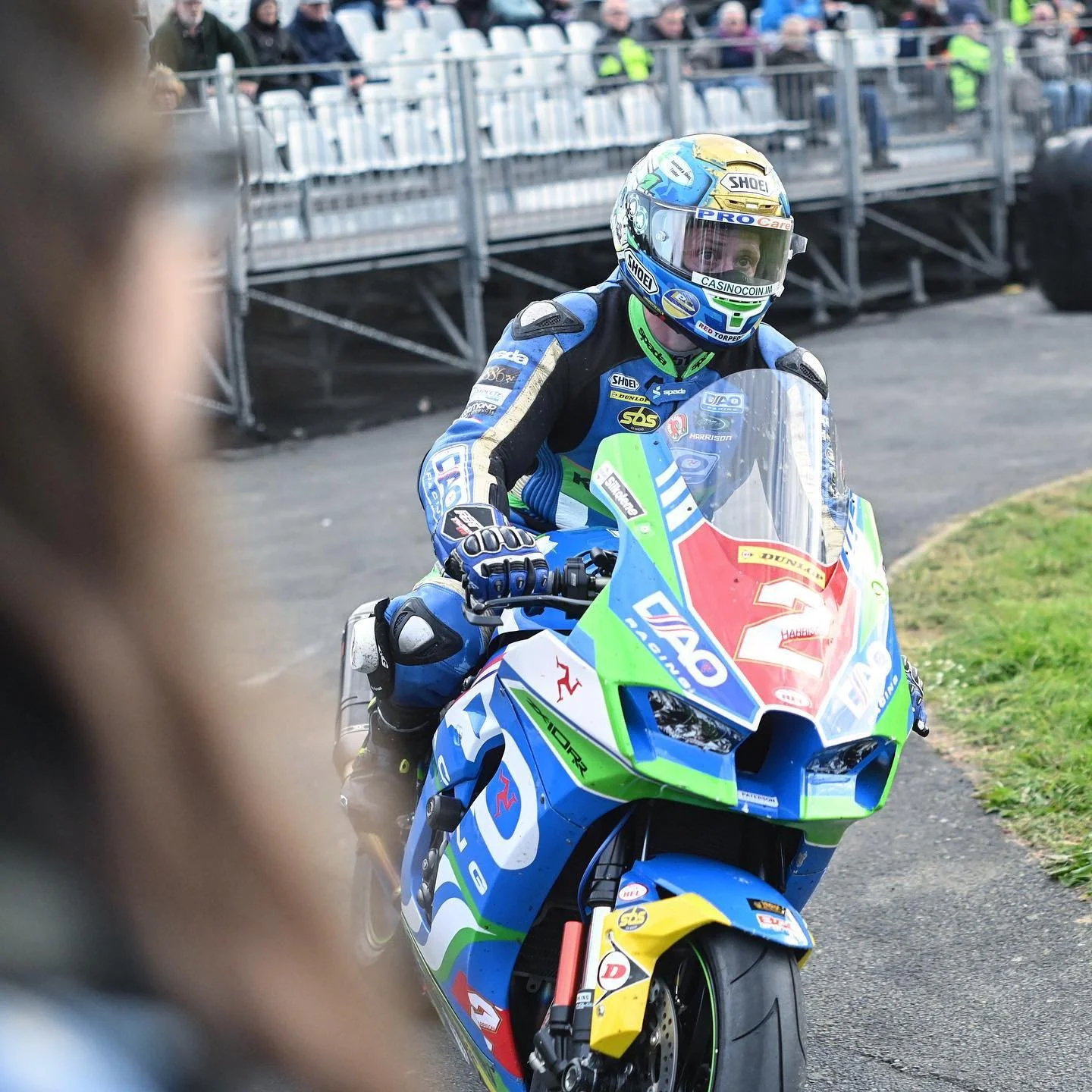 Race car driver in full racing gear riding a blue, green, red, and yellow motorcycle on race track with spectators in the background.