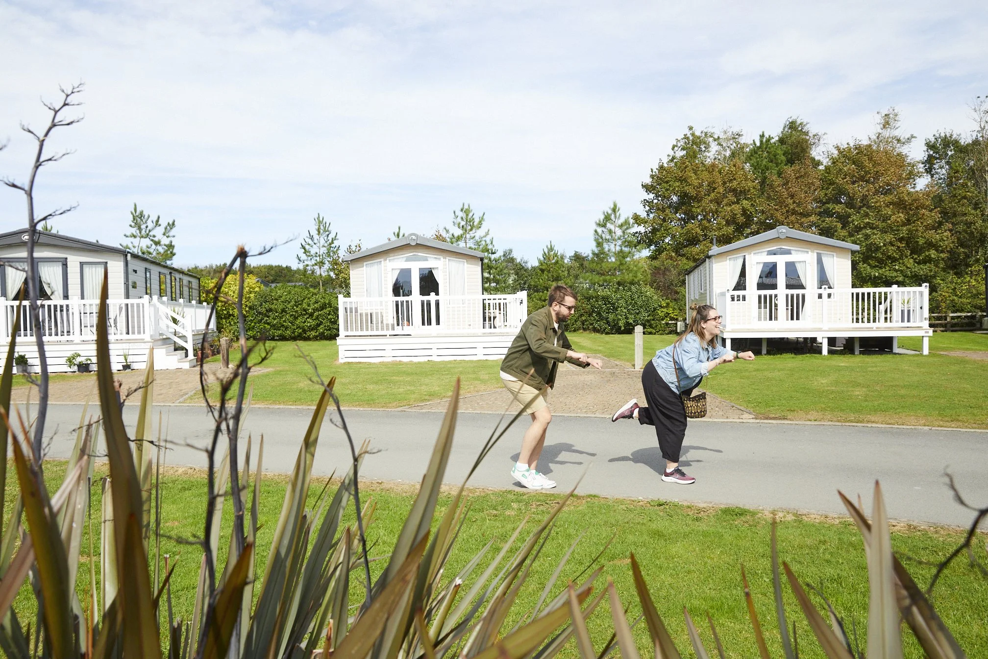 Two people pulling on a rope on a paved pathway in front of white cabins with decks, with grass, trees, and blue sky in the background.