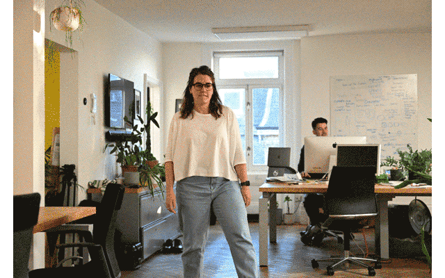 Woman standing in a bright, modern office space with desks, computers, and a person working at a desk in the background.