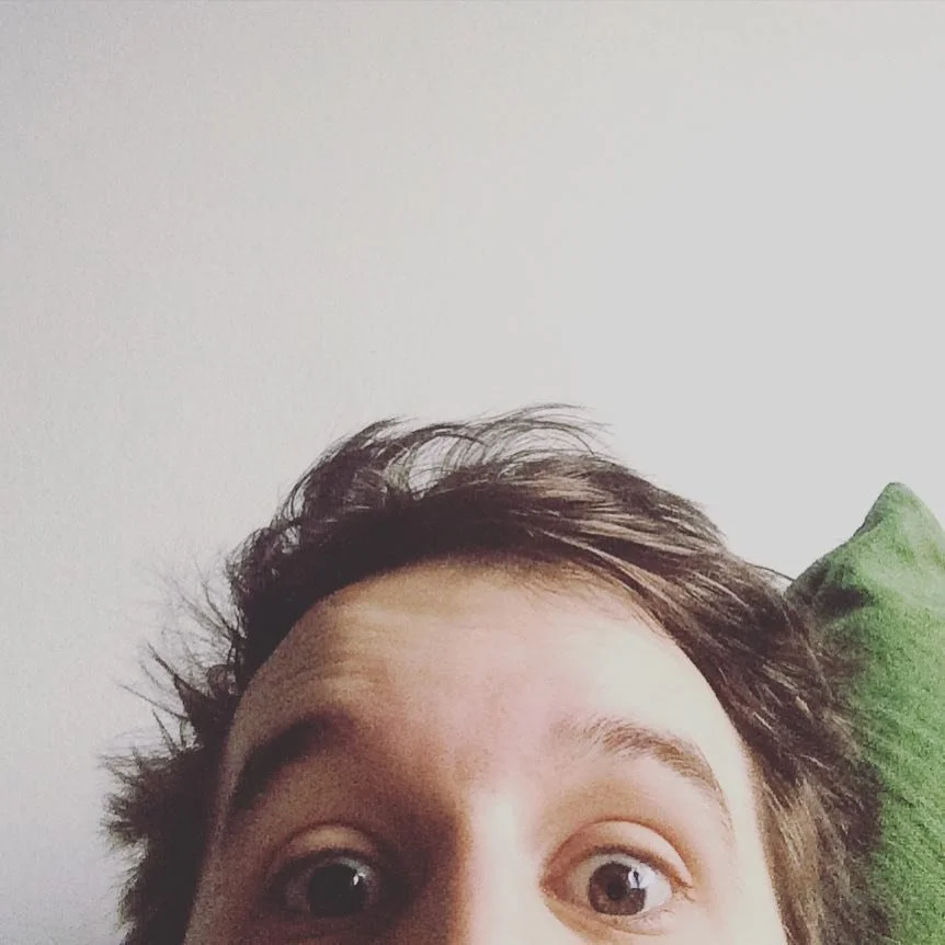 Close-up of a person with wide eyes and messy hair, looking at the camera, with a white wall and green pillow in the background.