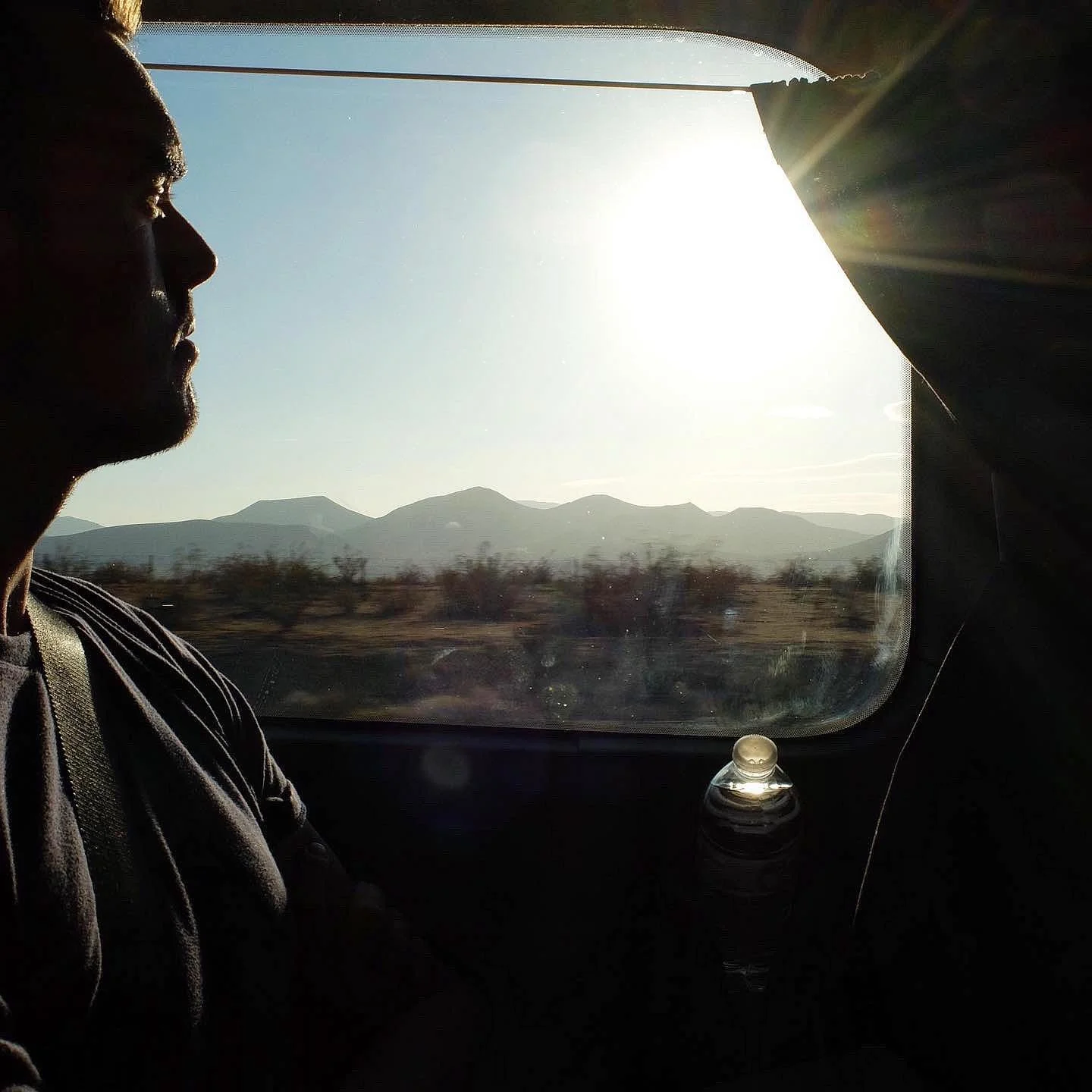 A person sitting next to a window during a sunset or sunrise, with mountains visible outside and a plastic water bottle on their lap.