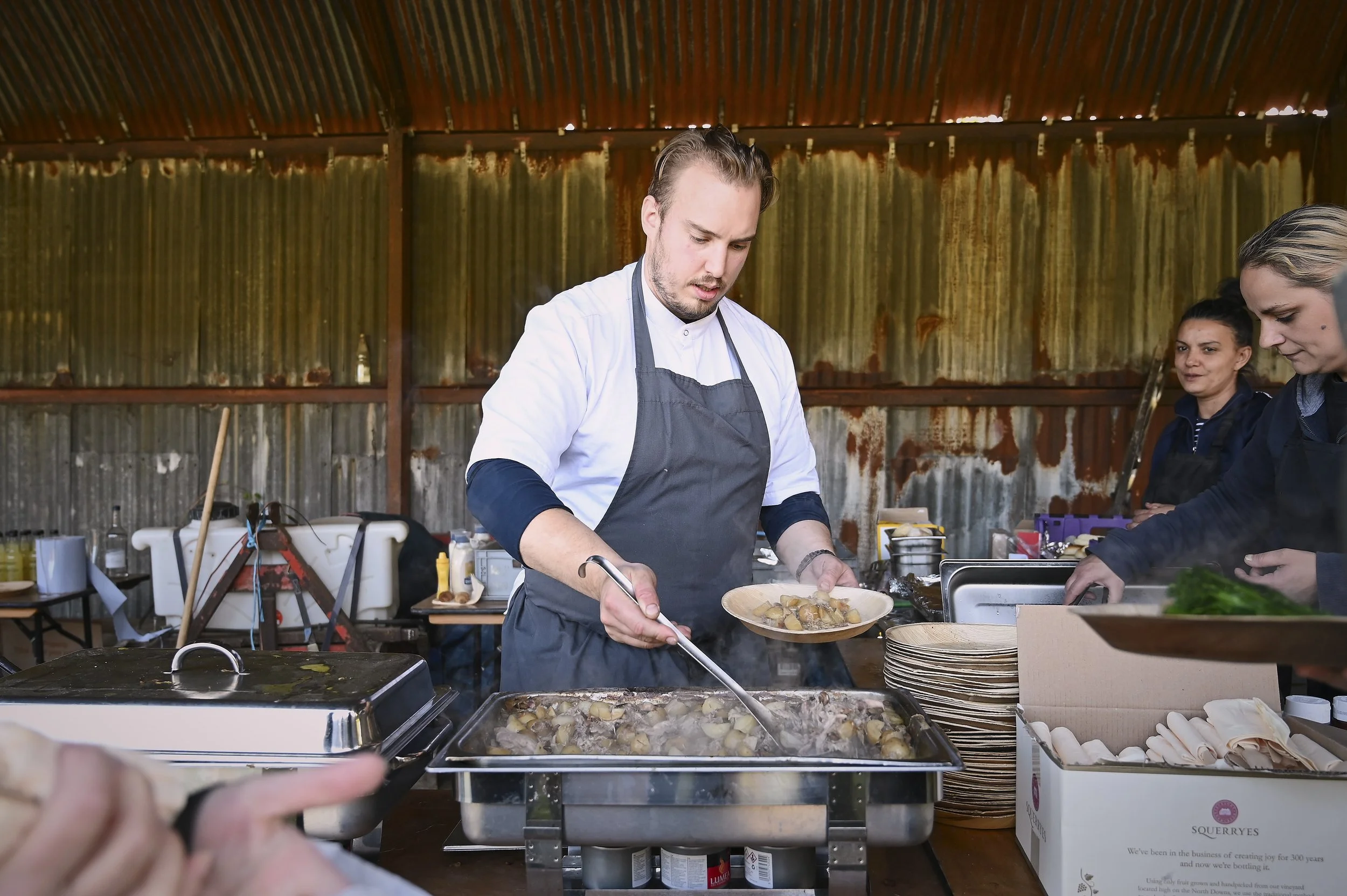 Chef serving food from a large tray in a rustic kitchen with women preparing dishes nearby.