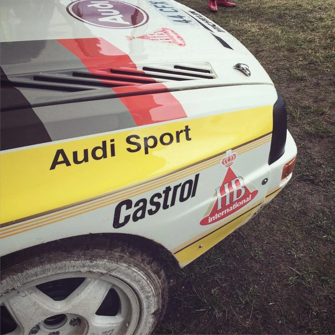 Close-up of a rally car with sponsor decals, including Audi Sport, Castrol, and HB International, showing part of the front wheel and hood. The car appears to be on a dirt or gravel surface.