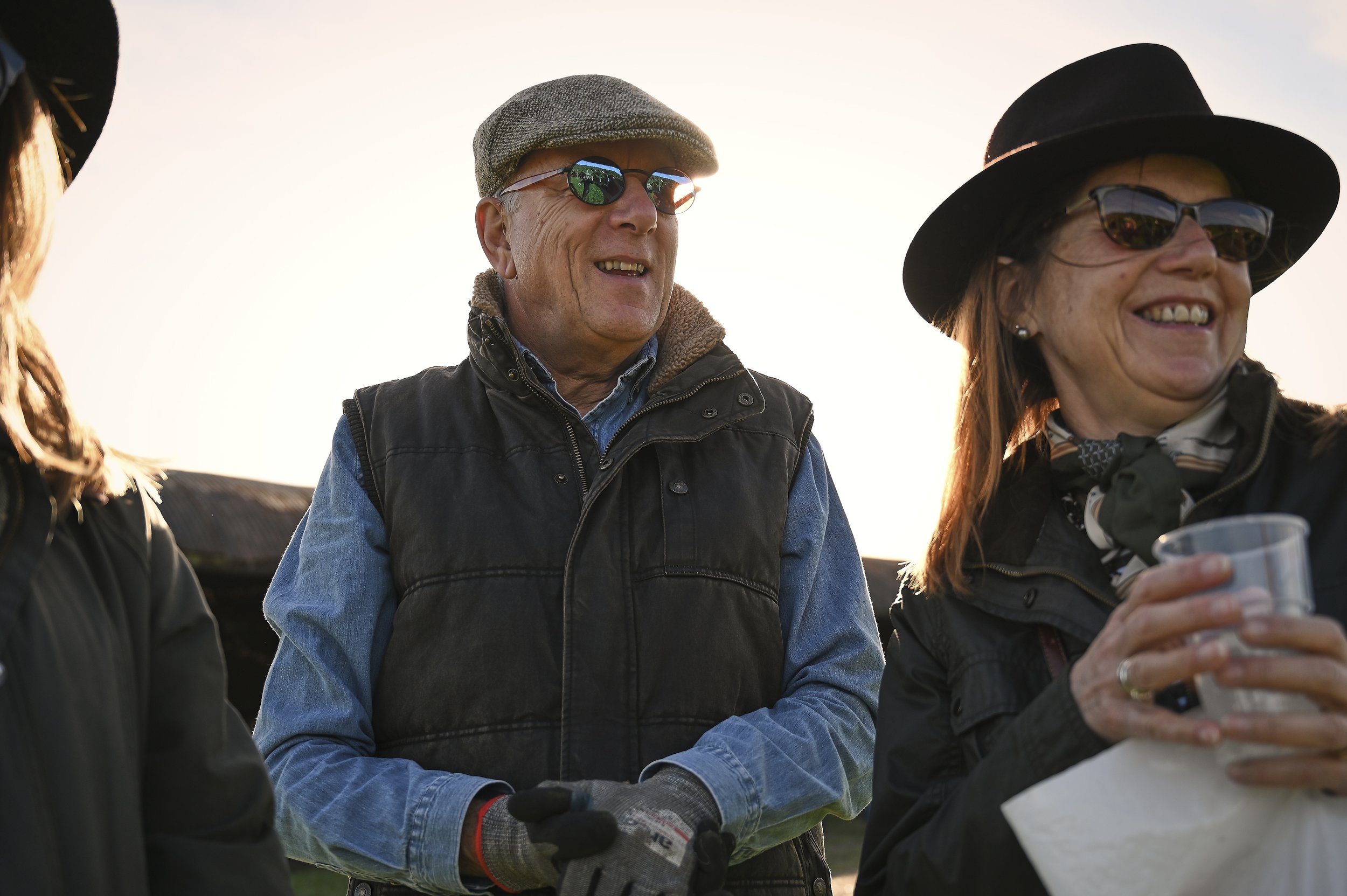 Group of three older adults outdoor at sunset, smiling and chatting; one woman holding a clear cup, wearing sunglasses and a wide-brimmed hat, two others wearing glasses and jackets.