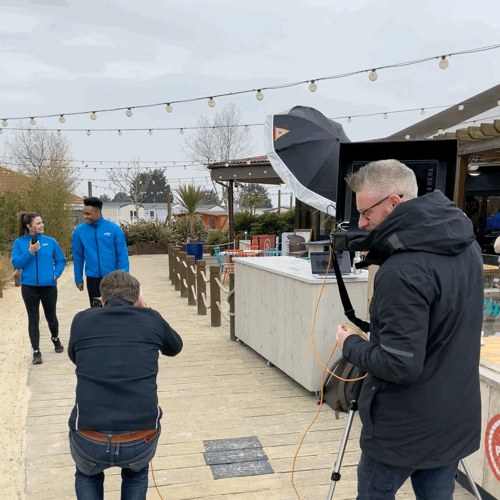 Filming crew captures footage of two people in blue jackets at outdoor venue with string lights and a wooden fence.