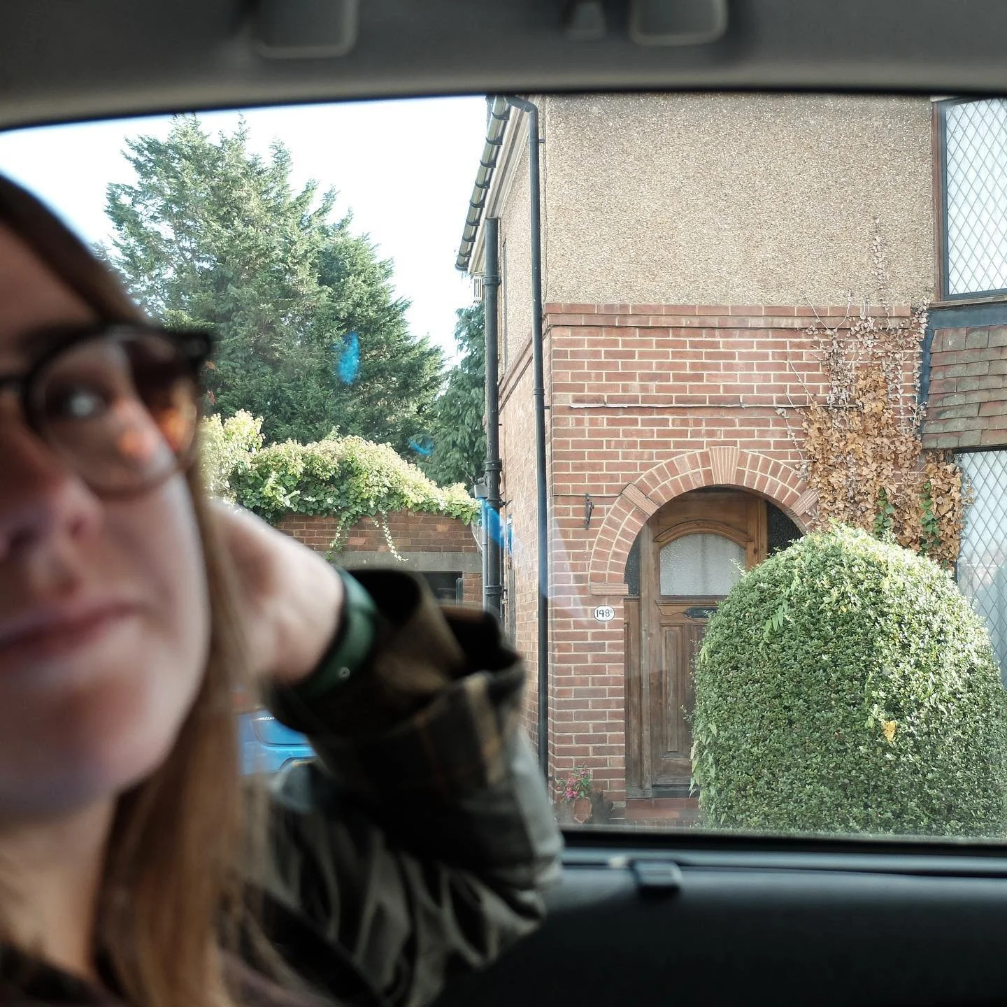 A woman with glasses and long hair sitting in a car, with a brick house, bushes, and trees visible through the windshield.