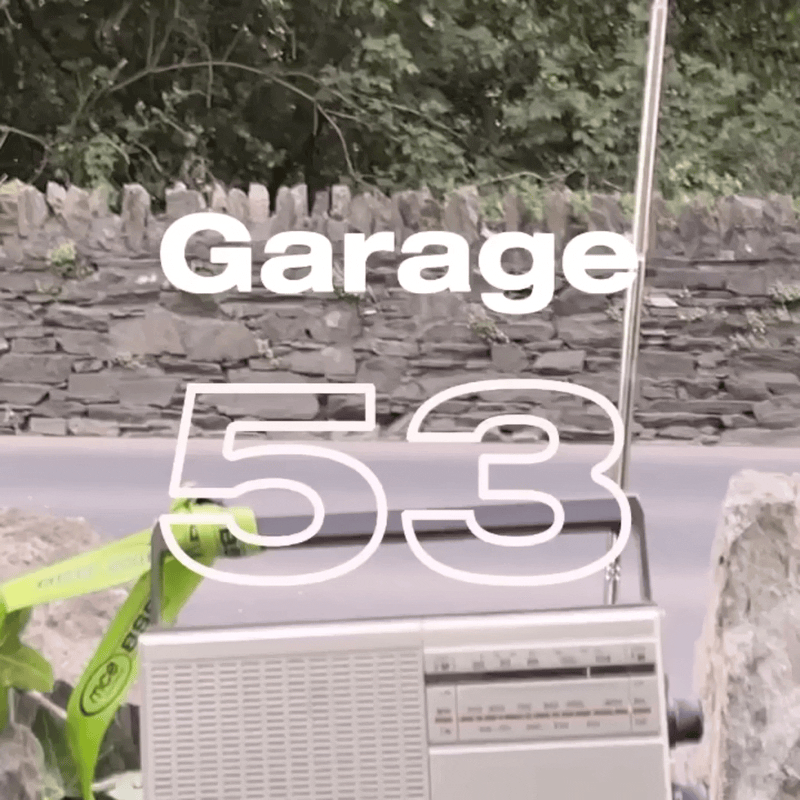 A street sign reading 'Garage 5-3' with a stone wall, greenery, and a gray street in the background, along with a portable radio or speaker in the foreground.