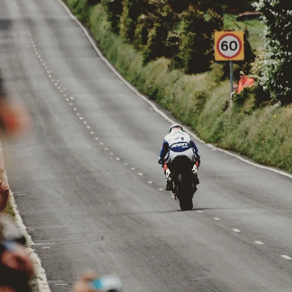 A motorcycle rider in a white and blue racing suit riding on a rural road with a 60 km/h speed limit sign on the roadside, surrounded by green foliage.
